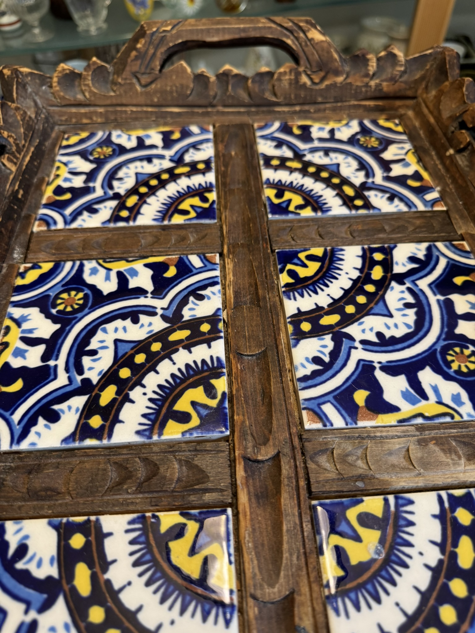 A close-up of a decorative wooden tray with four blue, yellow, and white patterned ceramic tiles. The tray has carved wooden edges, a handle at the top, and wooden dividers forming a cross shape over the tiles. The background is softly blurred.