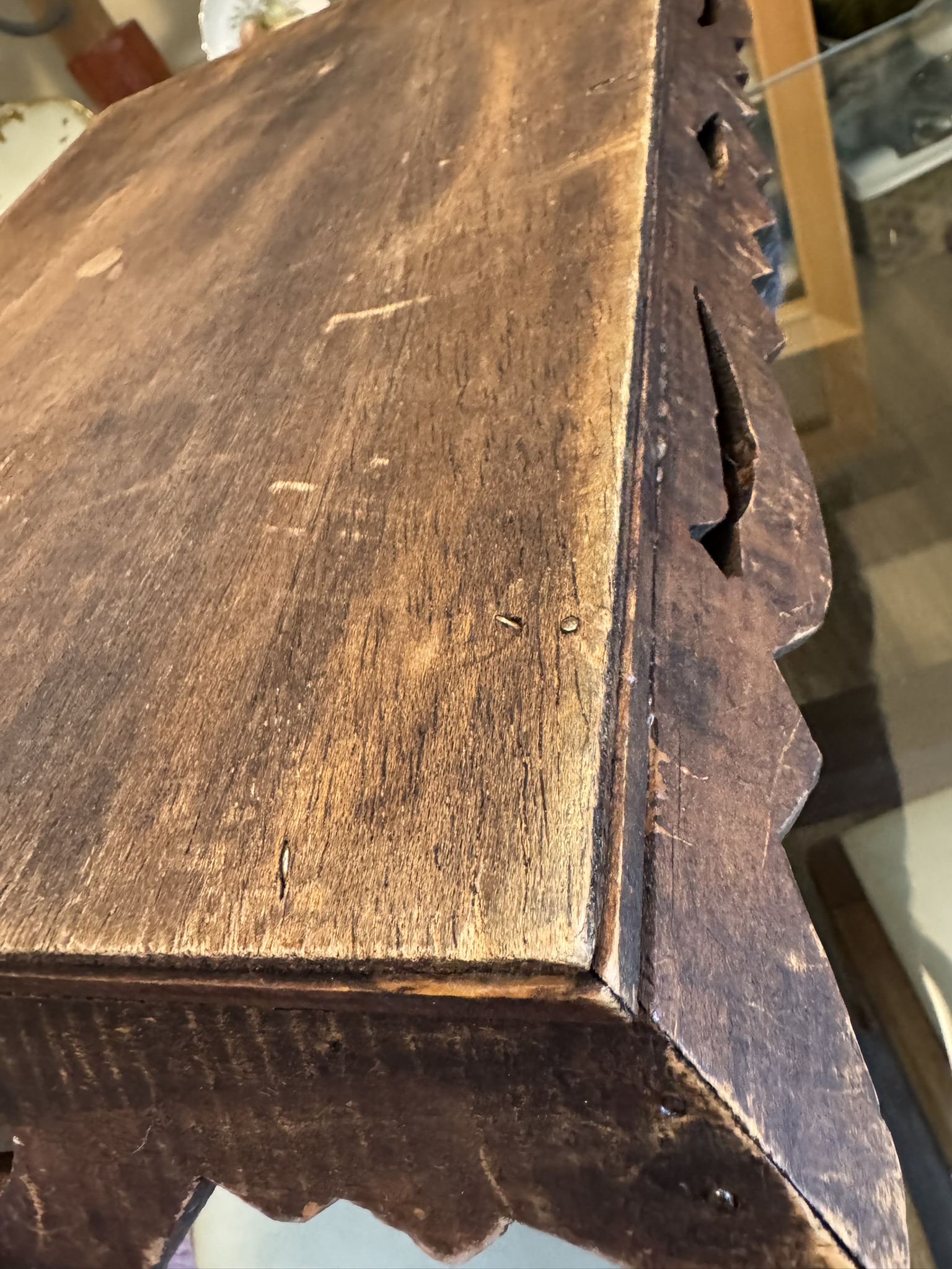 A close-up of a wooden table with a rustic, worn finish. The surface shows scratches and small nail heads. The edge features a scalloped or carved decorative trim. The photo captures the wood’s texture and aged appearance in warm lighting.
