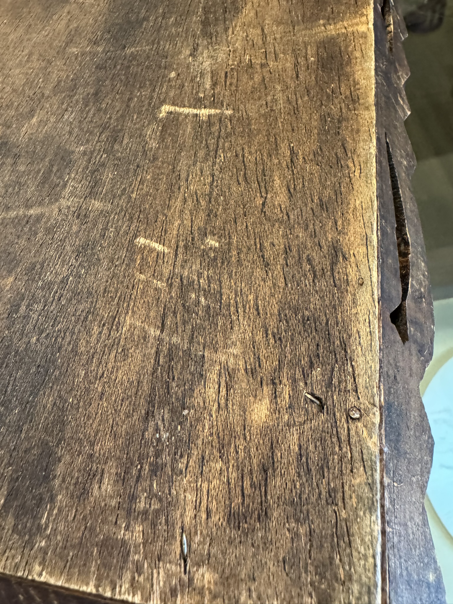 Close-up of a wooden tabletop with a rough texture, visible scratches, scuffs, and dents. The wood has a warm brown tone and uneven edges on the right side, with a jagged area. Several small nails and natural grain patterns are also visible.