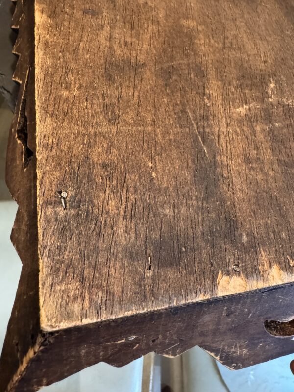 A close-up of an old, weathered wooden table with visible scratches, cracks, and uneven edges; the wood appears rough and faded, with a few small nails and notches, and the table rests on a tiled floor with sunlight casting a soft shadow.