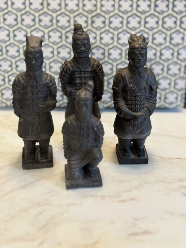 Four small dark statues of ancient Chinese terracotta warriors, three standing and one kneeling, are arranged on a light marble surface. The background has blue and white geometric tiles with an interlocking pattern.