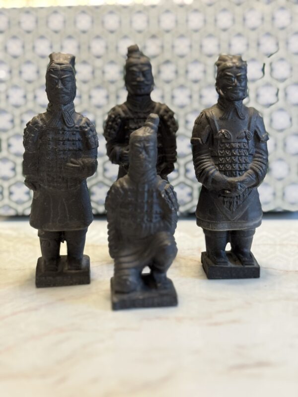 Four small, dark statues of Terracotta Warriors stand on a light surface with one figure kneeling. The background features a soft-focus blue and white geometric pattern, adding contrast to the detailed armor on the statues.