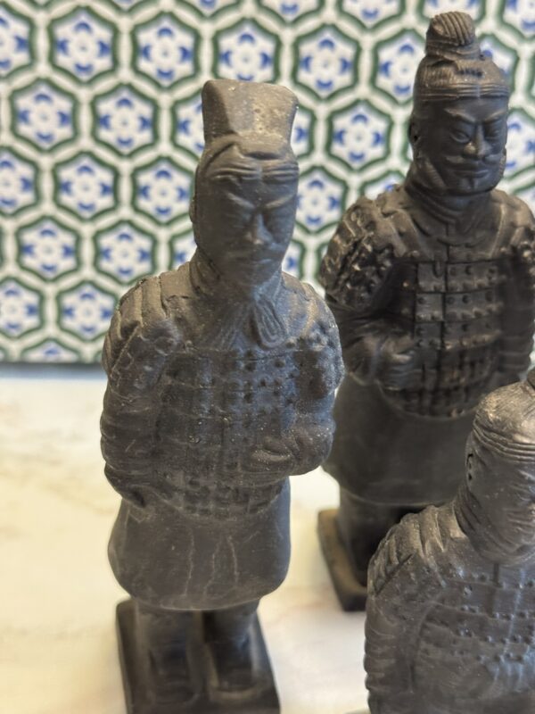 Three small, dark gray statues resembling ancient Chinese Terracotta Warriors stand on a light surface. The background features a blue, green, and white geometric tile pattern. The warriors wear detailed armor and have distinct headpieces.