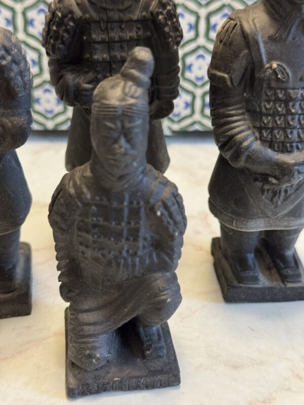 A close-up of four small, dark statues resembling ancient Terracotta Warriors. The front statue kneels on one knee, while the others stand. The background is a patterned tile with green, blue, and white geometric designs.