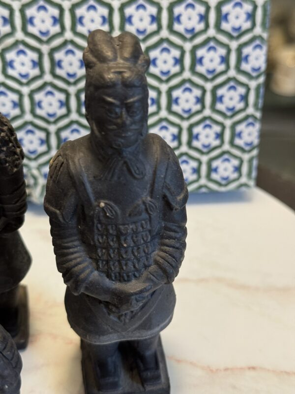 A small black figurine of a warrior with detailed armor and a helmet stands on a smooth, light surface. The background features a blue, green, and white geometric tile pattern. The figurine’s hands are clasped in front.