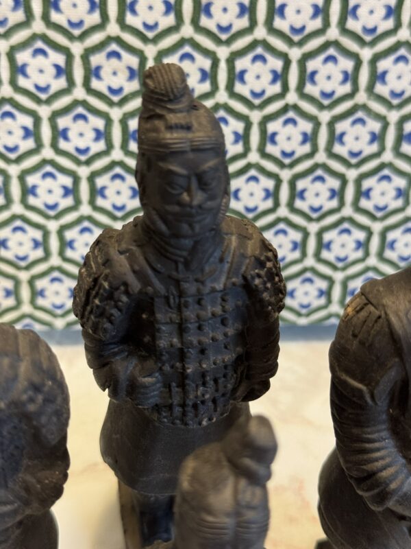 A close-up of a small, dark statue resembling a Terracotta Warrior, with detailed armor and a topknot hairstyle. In the background, there is a patterned blue, green, and white geometric tile. Other similar statues are partially visible.