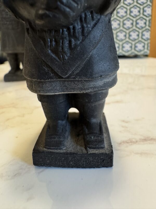 Close-up of a dark, weathered figurine standing on a square base. The figure has short legs and wears a coat with a triangular hem. The background shows a patterned blue-and-white tile and part of another similar figurine. Marble surface visible below.