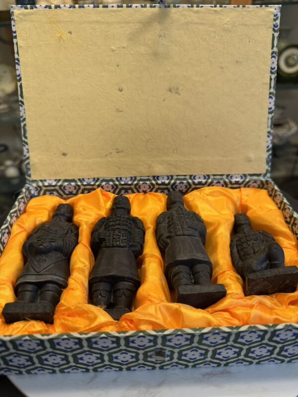 Four miniature terracotta warrior figurines lying in a decorative box lined with yellow satin fabric. The box has a geometric patterned exterior and an open lid. The warriors are dark in color and each has distinct armor and headgear details.
