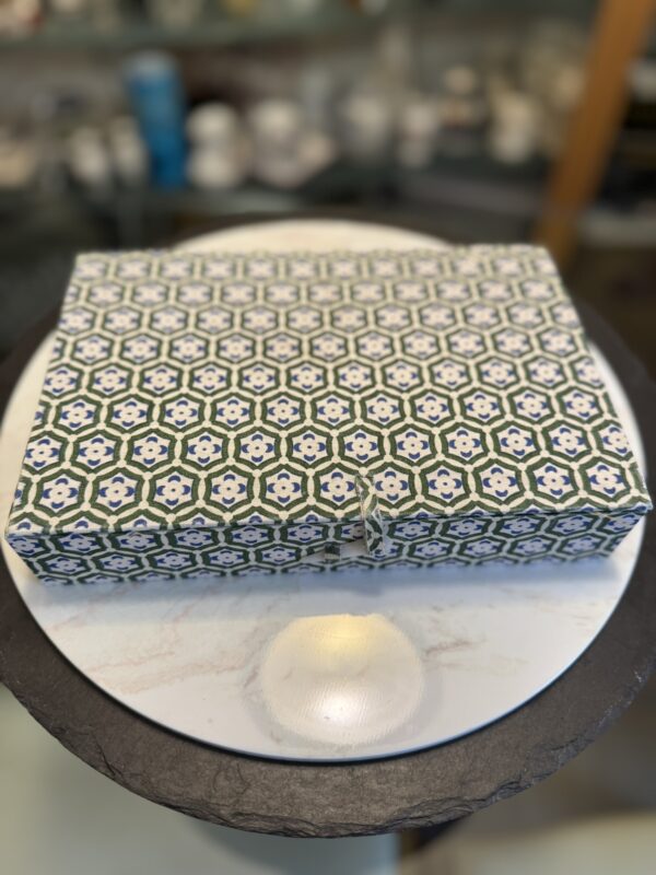 A rectangular box wrapped in geometric-patterned paper with green, blue, and white hexagonal designs sits centered on a round, white marble-topped table. The background is softly blurred, showing shelves with various objects.