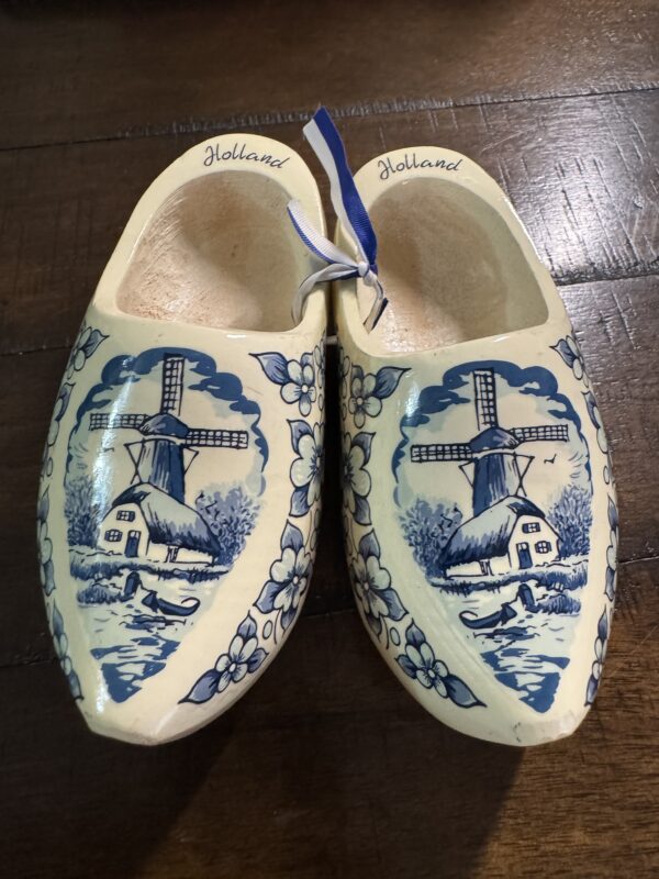Wooden Delft Clogs, marked "Holland," feature blue windmill and floral designs and are displayed on a dark wood surface. A blue-and-white ribbon ties the decorative clogs together.