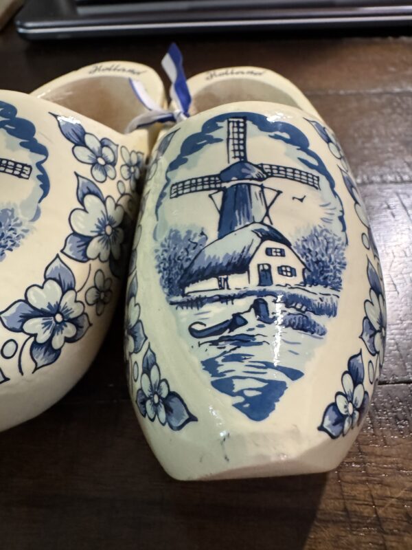 A decorative pair of Wooden Delft Clogs painted blue and white: one features a windmill, house, trees, bird, and florals; the other shows part of a floral design. Both wooden clogs are displayed on a wood surface.