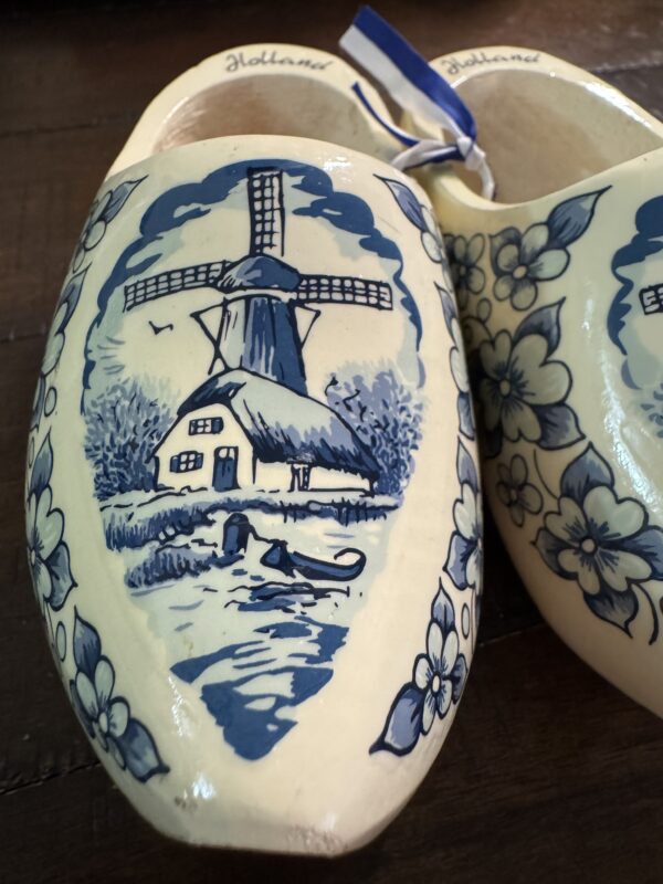 A close-up of Wooden Delft Clogs, cream-colored shoes with intricate blue floral designs and a windmill motif. 