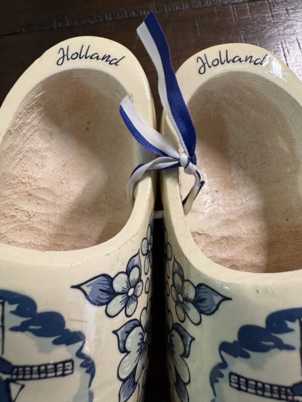 Wooden Delft Clogs: Pair of cream ceramic clogs featuring blue floral and windmill designs, “Holland” on each heel, and a blue-and-white ribbon, styled after traditional shoes.