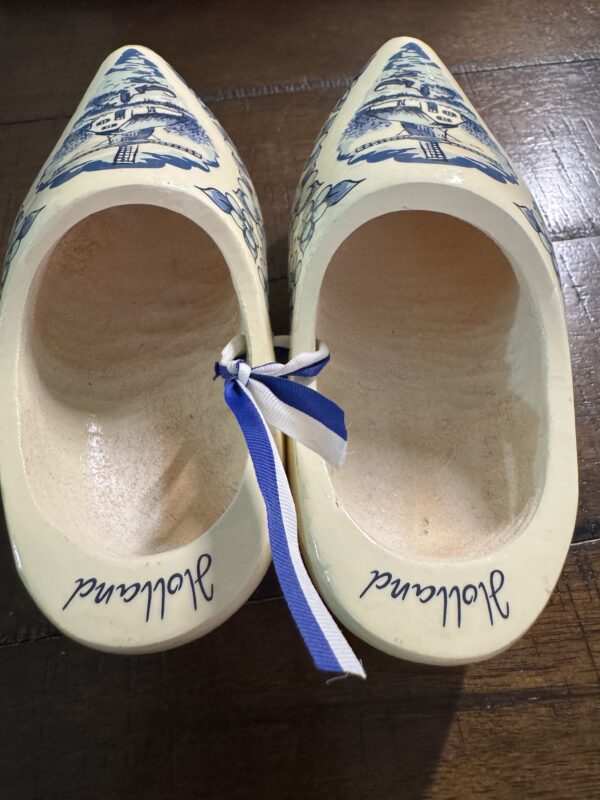 The Wooden Delft Clogs are cream-colored with blue windmill designs on the front, “Holland” marked inside in cursive, and a blue and white ribbon tying them together at the heels.