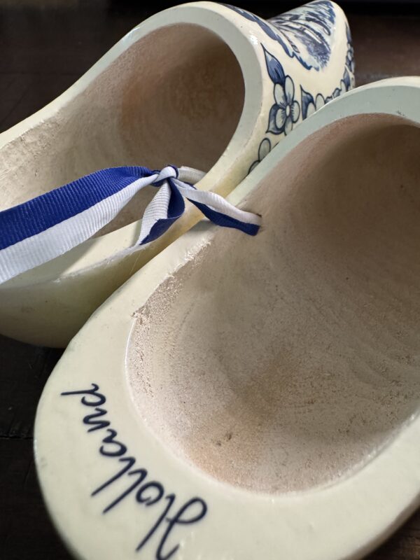A close-up of two cream-colored Wooden Delft Clogs with blue floral patterns. One clog has a blue-and-white ribbon, and 