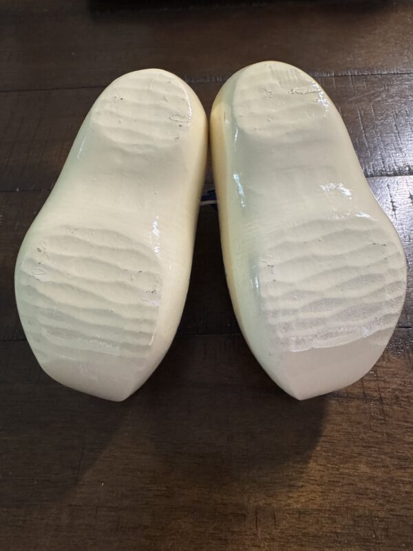 A close-up of the soles of two Wooden Delft Clogs in light beige, resting on a dark wooden surface. Their shiny, grooved soles reflect light, suggesting they are new or well-polished.