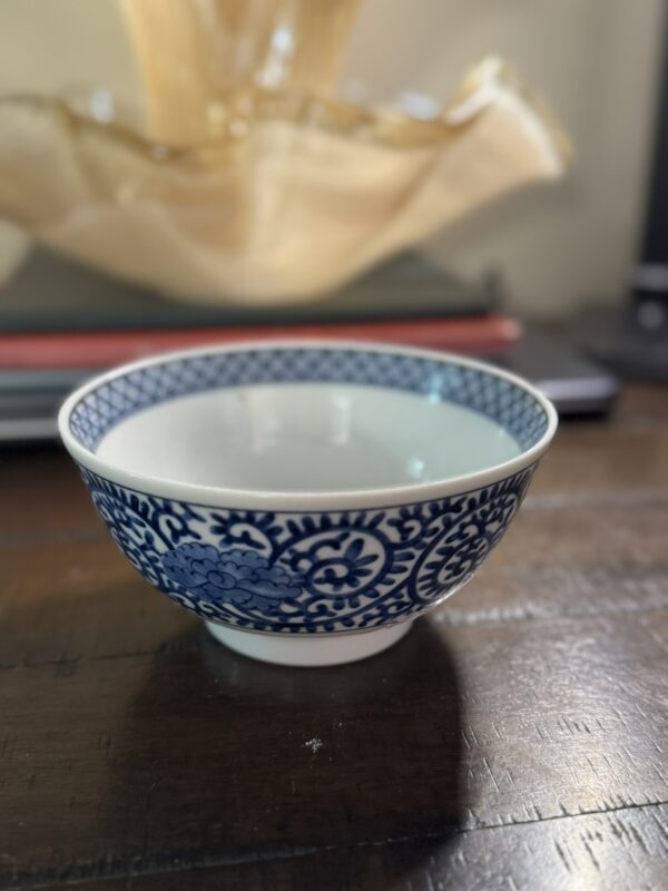 A small Tako Karakusa Bowl with detailed blue floral patterns sits on a wooden surface. In the background, a translucent beige decor item and a closed laptop with a red cover are softly blurred.