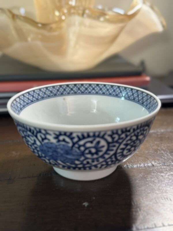 The Tako Karakusa Bowl, with blue floral and geometric patterns on white porcelain, rests on a dark wooden surface. Its interior rim displays a blue patterned border, while a glass decor piece and stacked items appear blurred in the background.