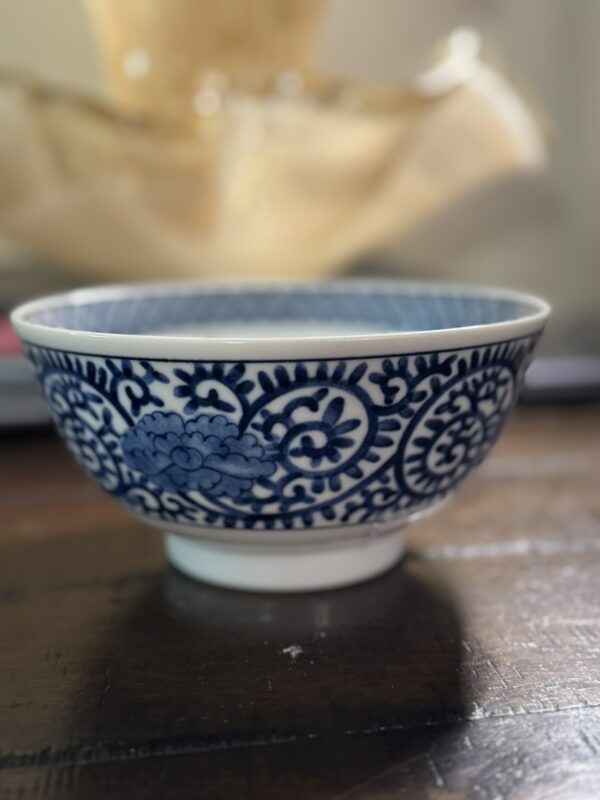 The Tako Karakusa Bowl, featuring blue floral and vine patterns on white porcelain, rests on a dark wooden surface with a softly blurred, warm cream background and gentle lighting.