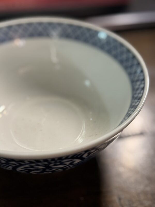 A close-up of the Tako Karakusa Bowl, empty and white with a blue geometric rim pattern, sits on a wooden surface. The image softly blurs at the edges, accentuating the bowl’s distinctive decorative border.