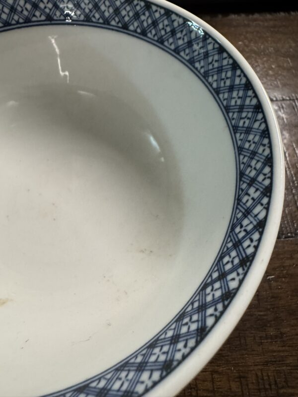 The Tako Karakusa Bowl is a white ceramic piece featuring a blue geometric crisscross pattern along the rim, shown in close-up on a wooden surface with minor discoloration or staining visible inside.