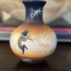 A small, round vase with a narrow neck features Southwestern-style art in earthy tones. Black figures, including a flute player and lizards, are painted with white and orange gradients and swirling patterns. The vase sits on a dark wooden surface.