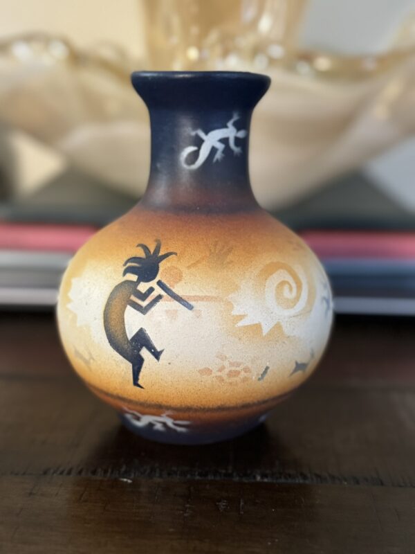 A small, round vase with a narrow neck features Southwestern-style art in earthy tones. Black figures, including a flute player and lizards, are painted with white and orange gradients and swirling patterns. The vase sits on a dark wooden surface.