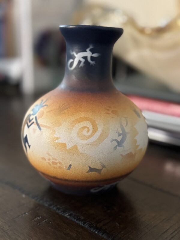 A round, narrow-necked clay vase features desert tones of orange, brown, and blue with painted designs of lizards, a spiral, and stick-figure humans inspired by Southwestern or Native American art, sitting on a dark wooden surface indoors.