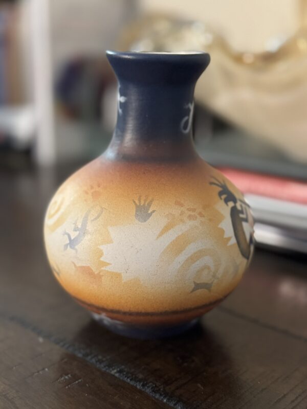 A small, round ceramic vase with a narrow neck, featuring a gradient from dark blue at the top to earthy orange and beige tones below. The vase is decorated with Native American-style motifs, including handprints, lizards, and swirling patterns.