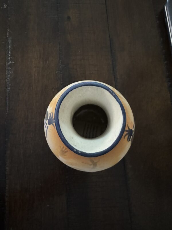 A small, round ceramic vase with a wide opening sits on a dark wooden surface. The vase is beige with blue trim and features black decorative designs, possibly stylized animals or figures, around its rim and body. Light shines from above.