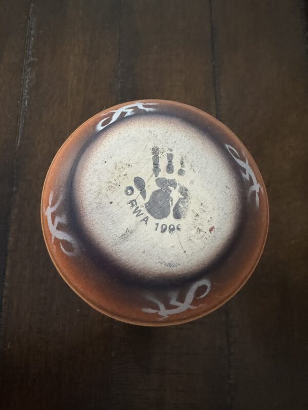 The bottom of a round, reddish-brown pottery piece with white curved designs. At the center, a black handprint is stamped with the text “ONWA 1996” beneath it. The pottery sits on a dark wooden surface.