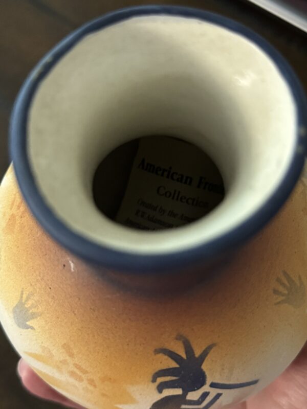 A hand holds a small ceramic vase with a blue rim and orange-yellow gradient. Stylized figures are painted on the vase. Inside, a label partly reading “American Frontier Collection” is visible at the bottom.