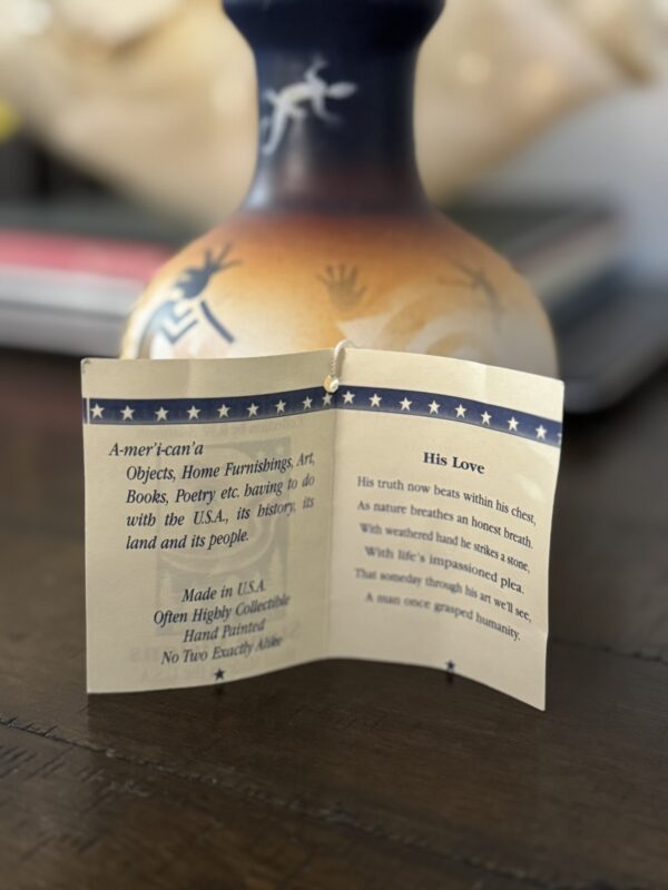 A small open booklet stands upright, displaying two printed pages. The left page defines American and mentions items made in the USA, while the right page features a poem titled His Love. A blurred, blue-and-beige vase appears in the background.