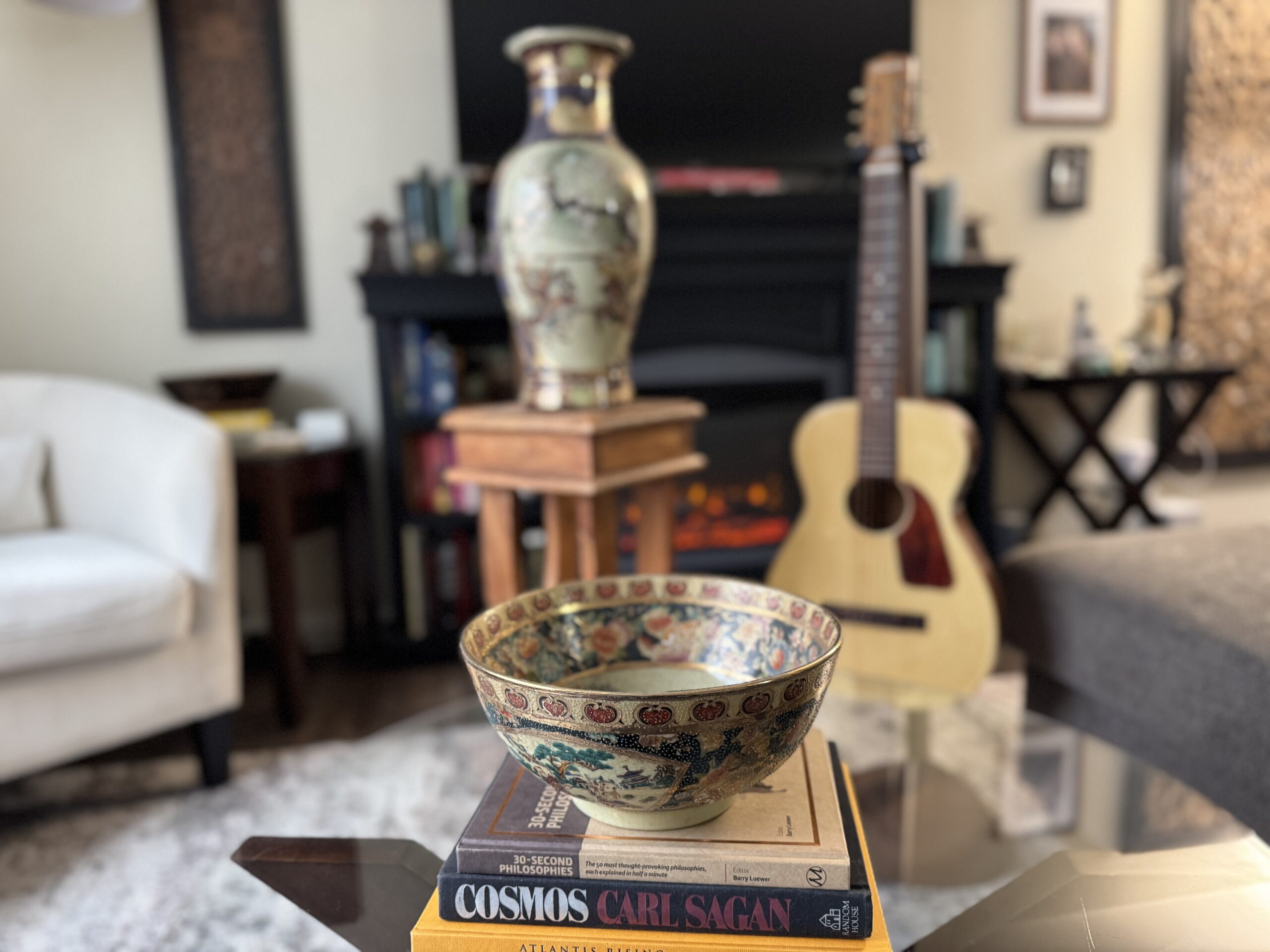 A decorative ceramic bowl with floral patterns—a touch of vintage decor—sits atop two books, including Cosmos by Carl Sagan, on a glass coffee table. In the background are a guitar, ornate vase, fireplace, and warm home furnishings.