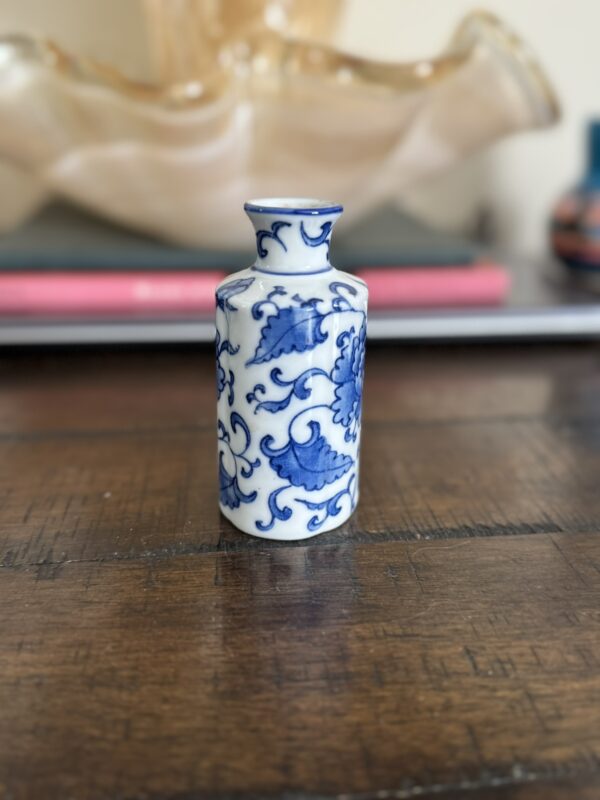 A small, white porcelain vase with intricate blue floral and vine designs sits on a wooden table. In the blurred background, there are some closed books and a beige, decorative glass object.