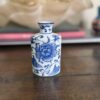 A small porcelain vase with a narrow neck features blue floral designs on a white background. It stands on a wooden surface, with blurred objects and a large decorative item in the background. The vase’s design suggests an Asian influence.