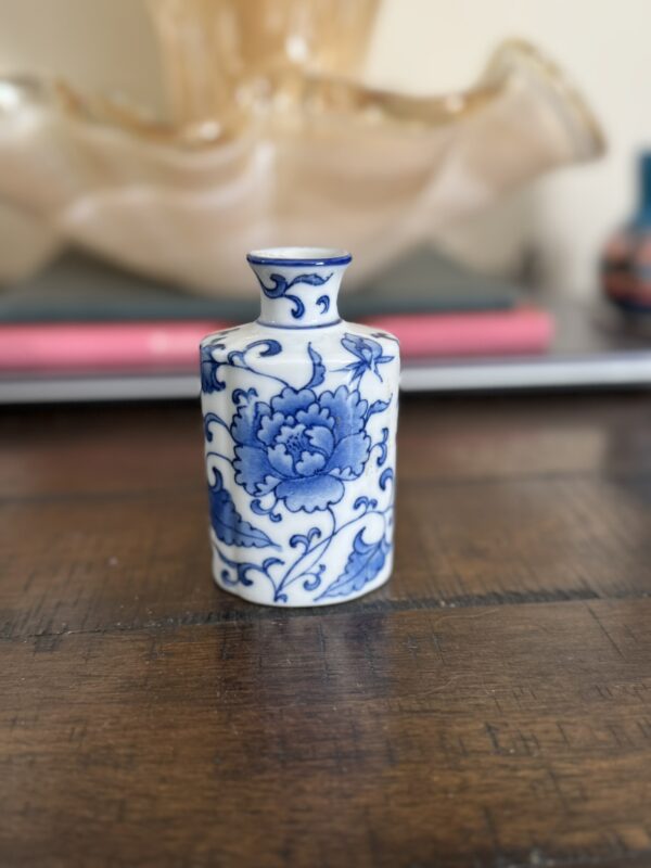 A small porcelain vase with a narrow neck features blue floral designs on a white background. It stands on a wooden surface, with blurred objects and a large decorative item in the background. The vase’s design suggests an Asian influence.