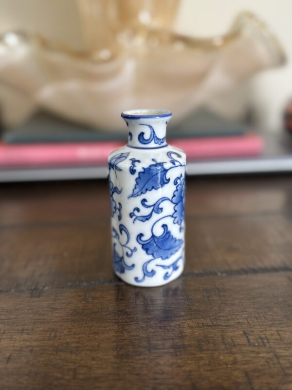 A small white porcelain bottle with blue floral and leaf patterns stands on a wooden surface. The background is softly blurred, showing a pink object and a beige decorative item, emphasizing the bottles delicate design.